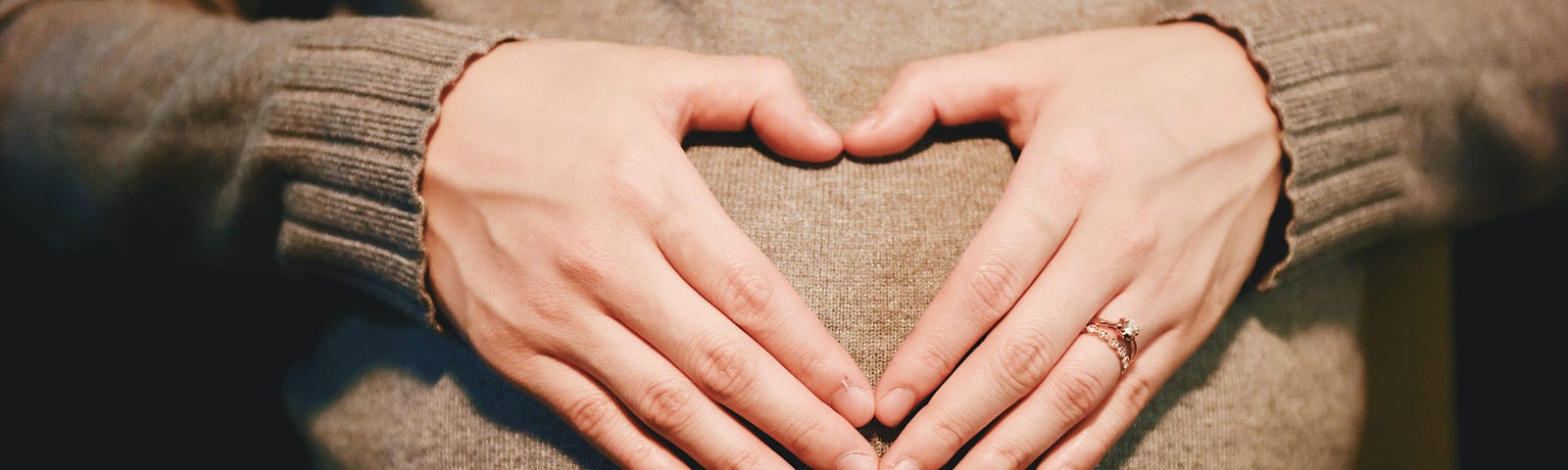 A pregnant woman in a brown sweater holder her hands in a heart shape over her belly.