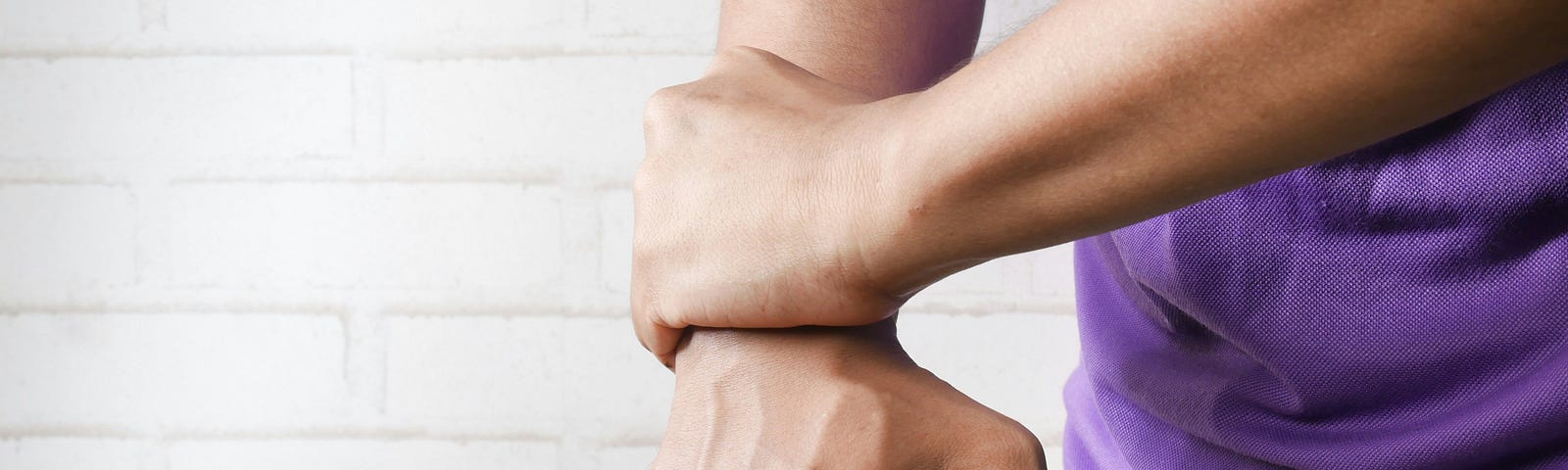 A man in a lavender shirt — you can’t see his face — holds on to his right wrist with his left hand.