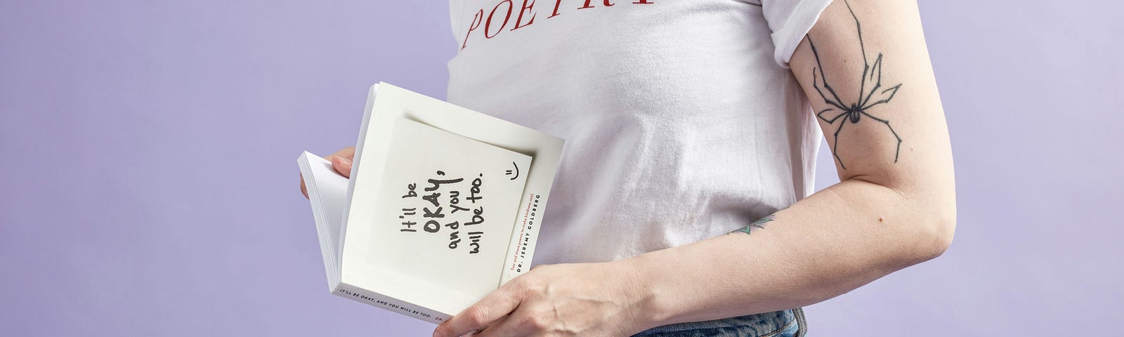 Person with a shirt that says Poetry, a spider tattoo on their arm and a book in their hand with what looks like a positive saying. Poetry?