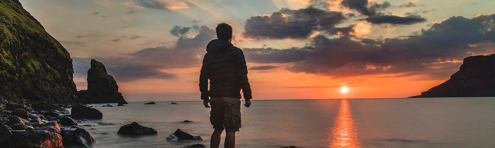 A man in shorts and a hoodie stands barefooted on a rock in a smoking sea, watching a fiery sun on the horizon, clouds above it, refracting the light like rays from a crown.