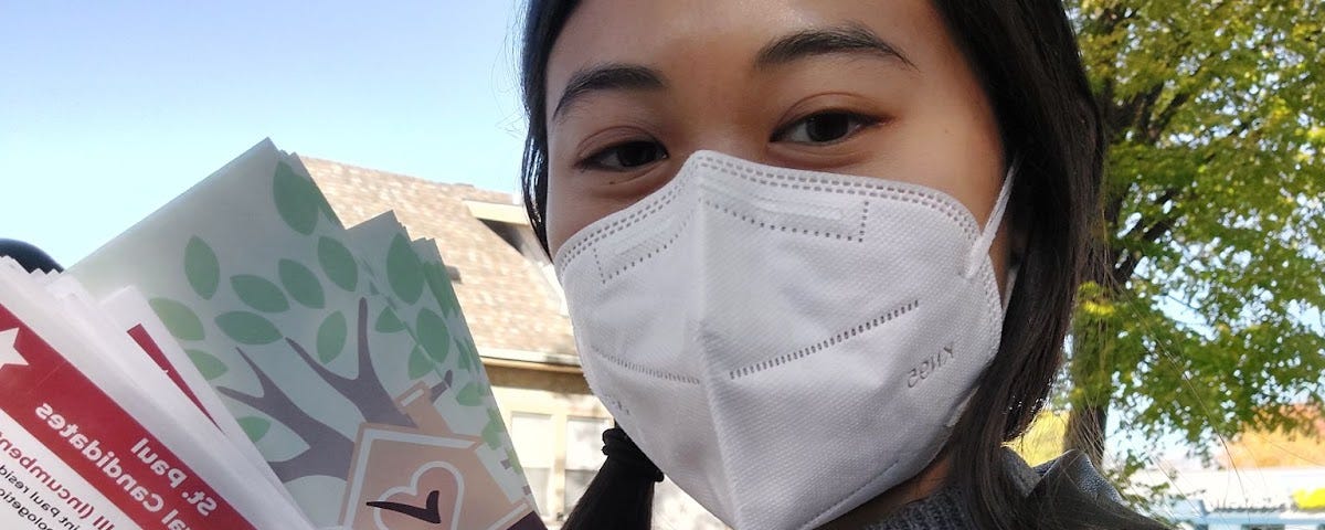 A photo of Leyen Trang wearing a face mask and holding up two sets of colorful fliers.