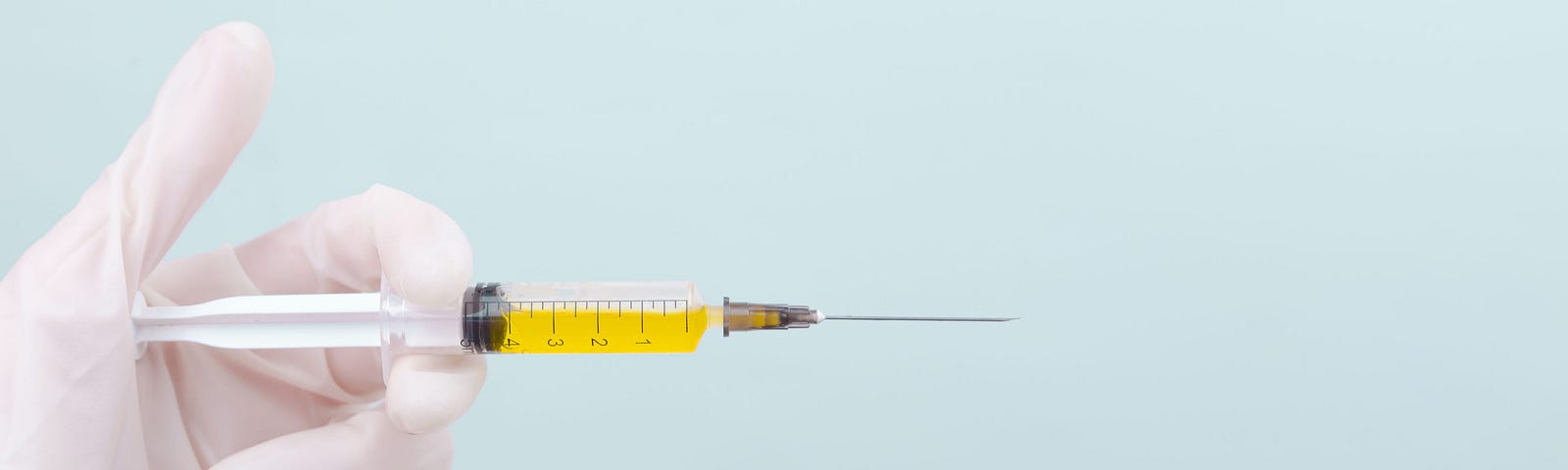 Photo of a gloved hand (at left of image) holding a syringe and needle. The syringe has yellow fluid and the hand is about to push on the plunger. Light blue background.