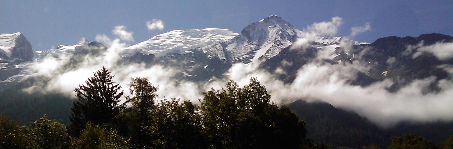 photo of snow-covered alps jutting up above some low clouds, with green trees and a tiny road sign in the foreground