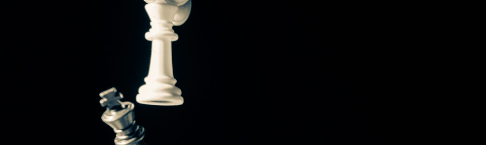 Hand from above holds white chess piece and is knocking over a black bishop piece. The image is in black and white and there are no other pieces on the board. Black background.