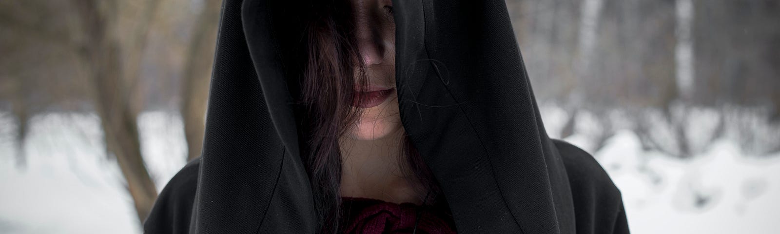 Close up hooded girl (dark brown hood) head bent, dark hair trailing at one side, eyes closed, head bowed. She wears a deep scarlet round-kneck dress with a silver talisman.