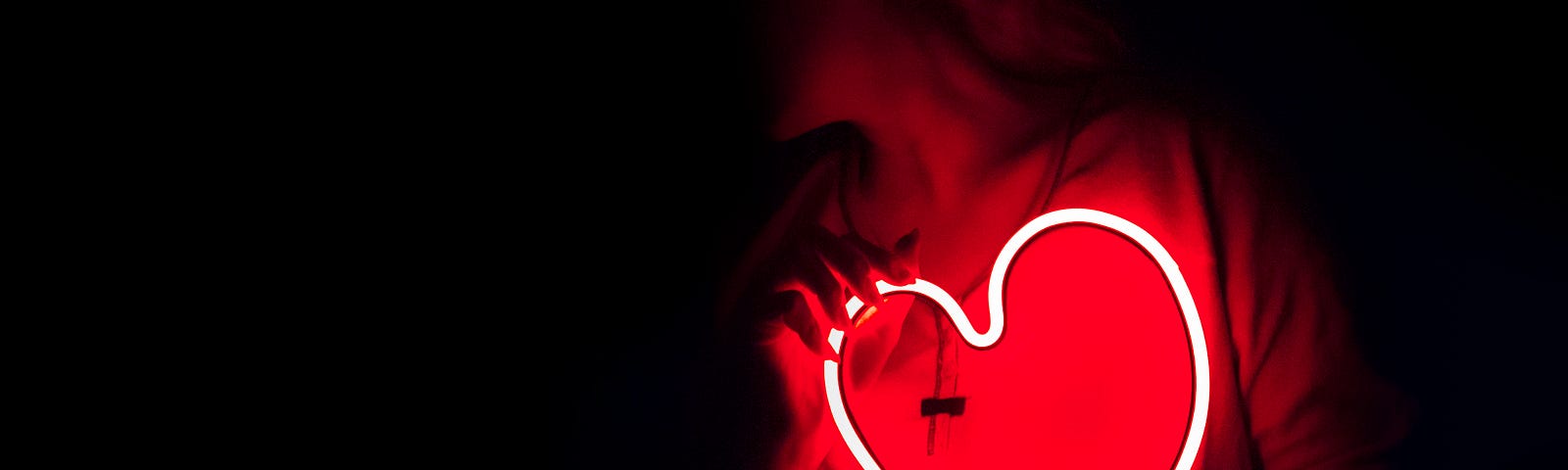 a woman holds a neon heart next to her red-sweatered chest. the rest of her is in shadow.