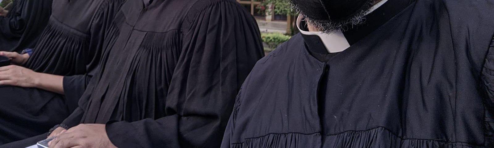 Três silhuetas de homens vestidos com batinas pretas e colarinhos brancos. Não mostra os rostos.