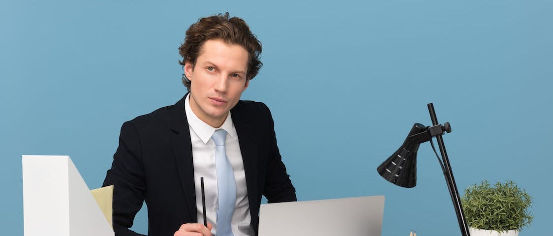 Man in black suit, white shirt, and pale blue tie sat at an office desk with a coffee, laptop, lamp, and other desk accessories in front of a blue wall, staring off into the distance thoughtfully
