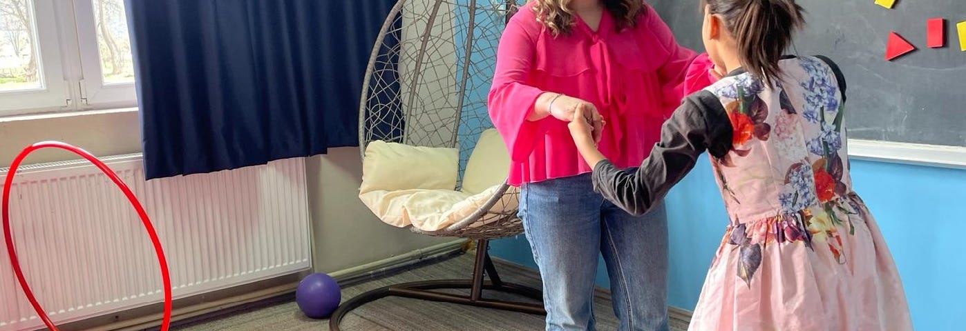 A 10-year-girl balances on a therapeutic board holding hands with a young woman standing across from her. There is a blackboard with magnets of different sizes and geometric shapes to their right. A self-standing hammock in the shape of an egg stands in the corner for swinging.