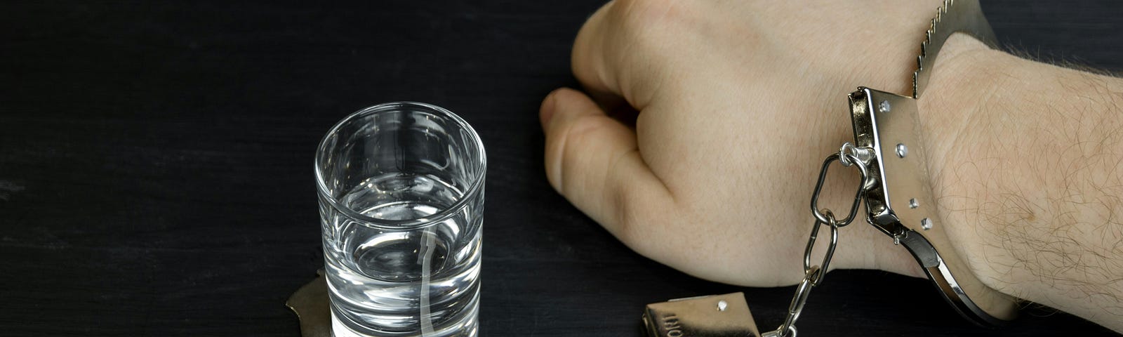 An image of a person’s hand, handcuffed to a shot of alcohol