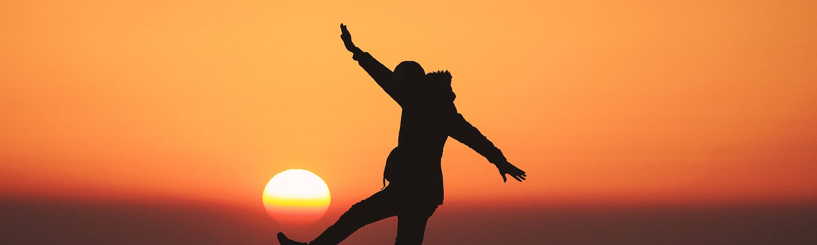 A person stands on their right foot on a rock. Their left lef extends forward at a 45 degree angle. The right hand extends upward for balance (and the left slightly downward). The sun is setting in the background, rendering the entire image an orange hue. One in five individuals in a recent study could not stand on one foot for ten seconds.