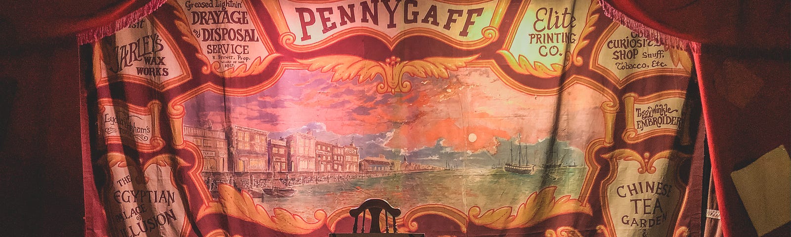 A stage with backdrop that says “PENNYGAFF” on it. A chair sits center stage with a box sitting on it.