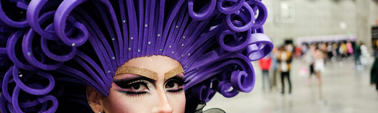 A drag performer with a bright purple wig looks at the camera
