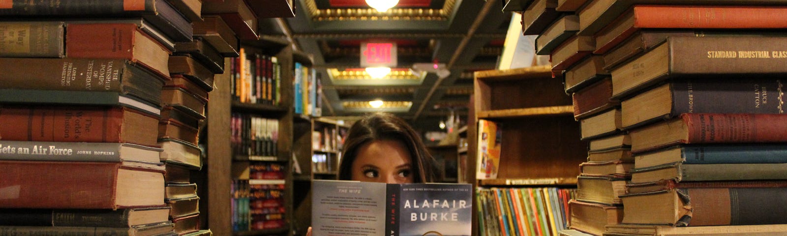 A woman reading a book while looking through a wall of books in a bookstore.
