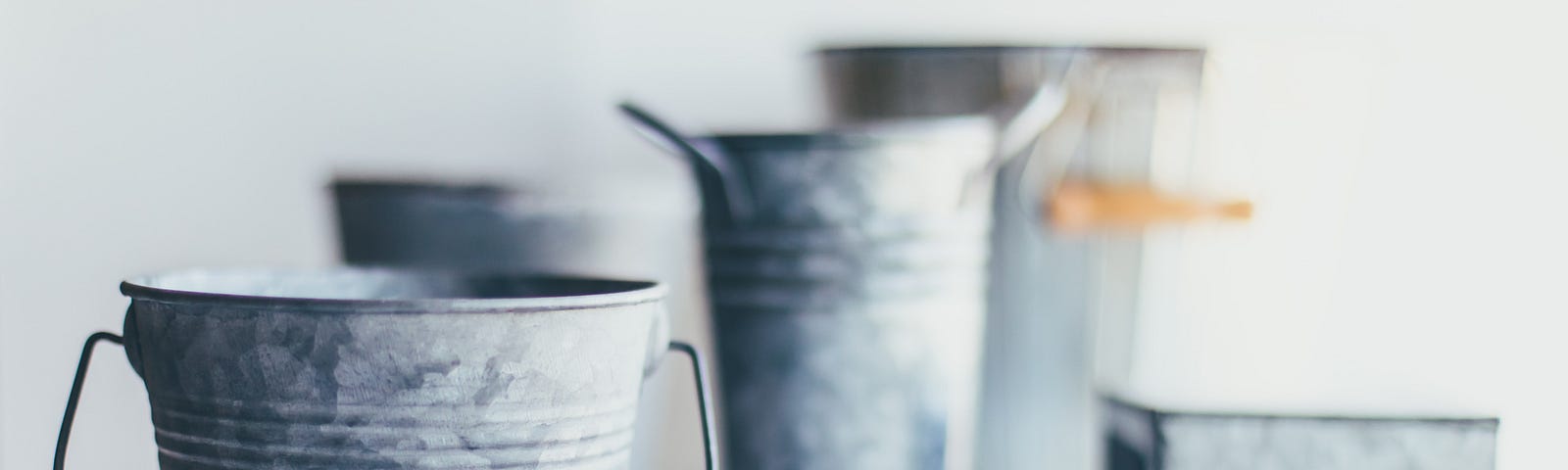 5 silver tin buckets. One with silver handles and two with silver handles with brown tracks. Standing on gray wooden floor.