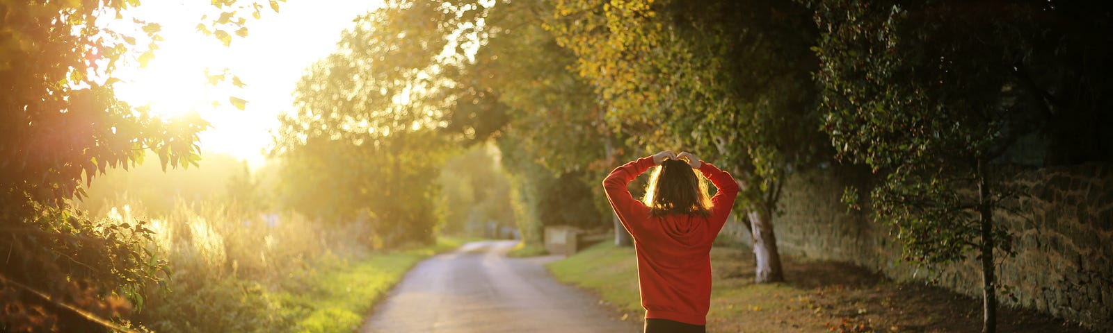 A woman walks on a path surrounded by trees, with the a bright white sky in the distance. The woman wears an orange sweatshirt and black sweatpants. Her arms are raised such that she has both hands on the top of her head.