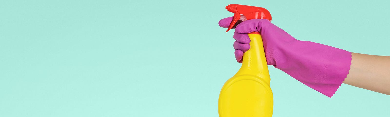 A pink gloved hand reaches across a light turquoise background, holding a yellow spray bottle