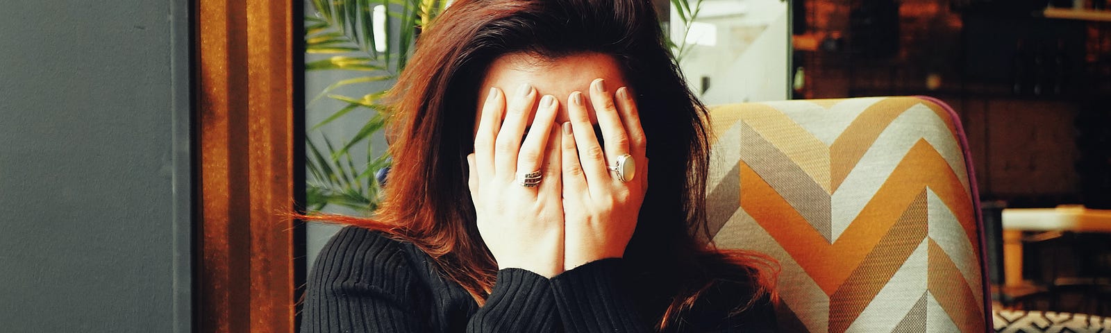 Woman with dark hair and black sweater sits in chair in front of window, covering her face with both hands.