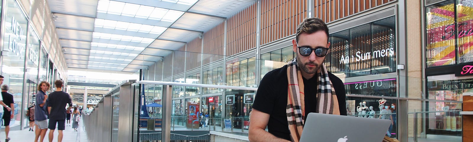 A man sitting at a mall with sunglasses while looking at his laptop