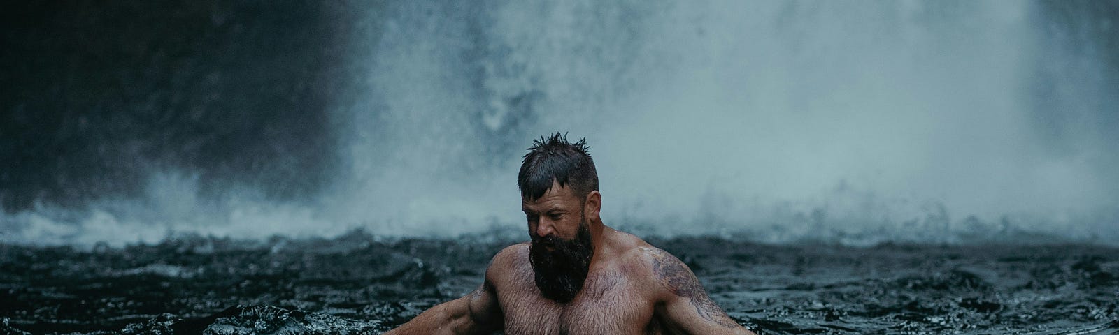 Shirtless strong man with a beard and tattoos in a body of water, with the bottom of a waterfall in the background. It’s raining, and his arms are outstretched into the water as he’s looking thoughtfully down at the choppy surface