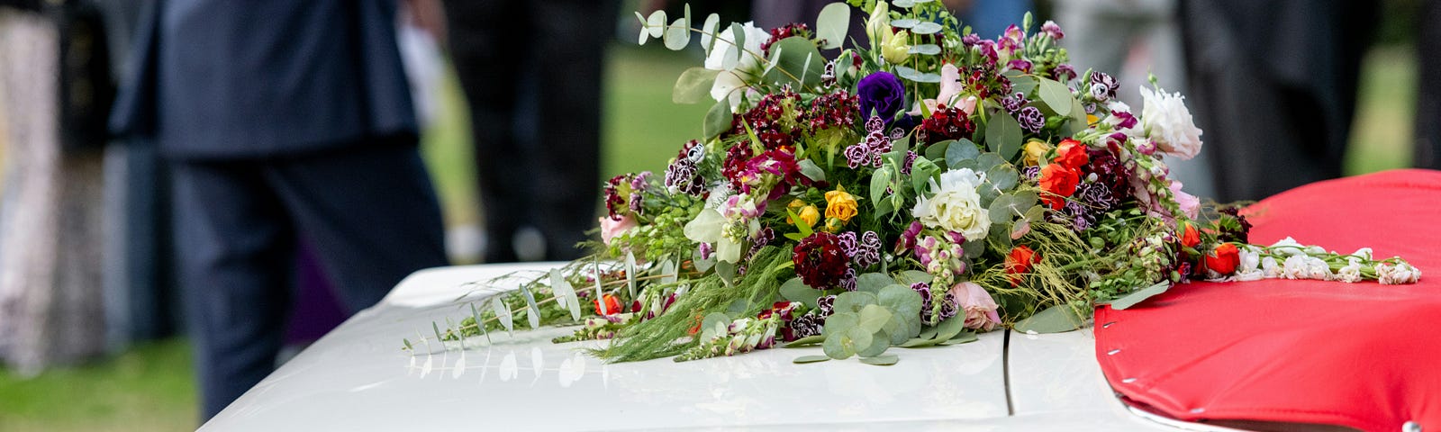 Photograph of a casket at a funeral. Floral arrangement on top. Crowd shown in background.