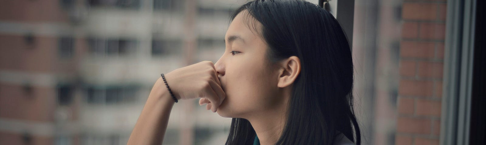 Young woman in gray sweat jacket, staring out window at highrise buildings, resting chin on hand, elbow on windowsill.