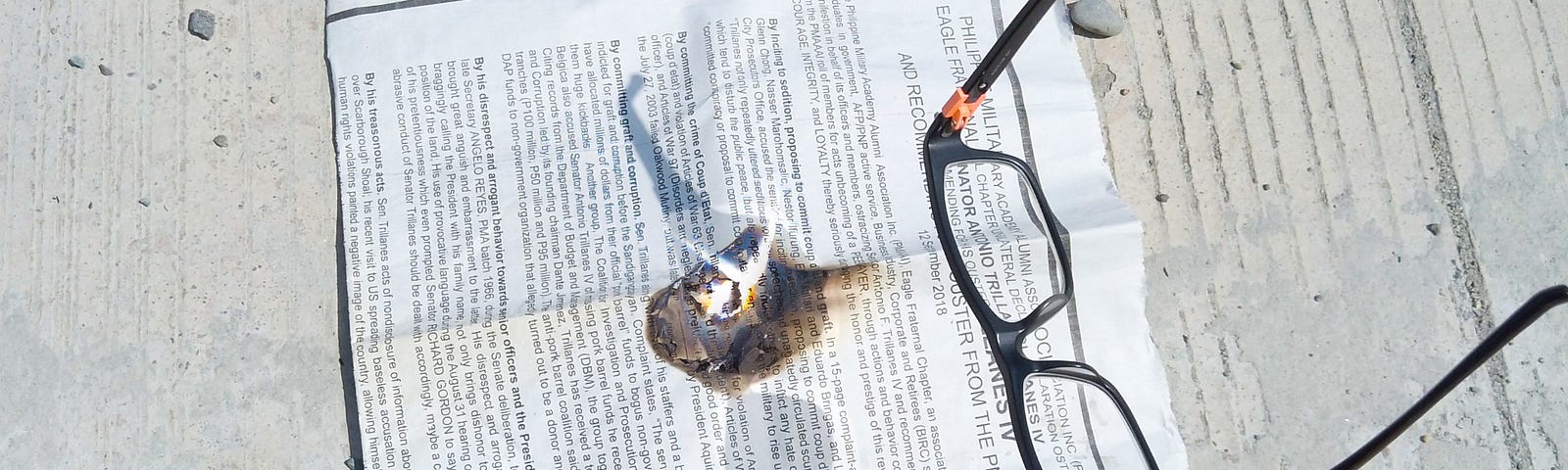 A pair of glasses are held under direct sunlight to start a fire on a newspaper.