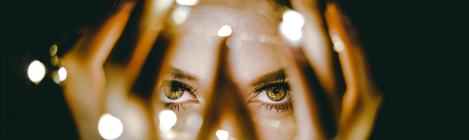 Woman in dark, reaching forward to hold strand of bright white lights. Black background.