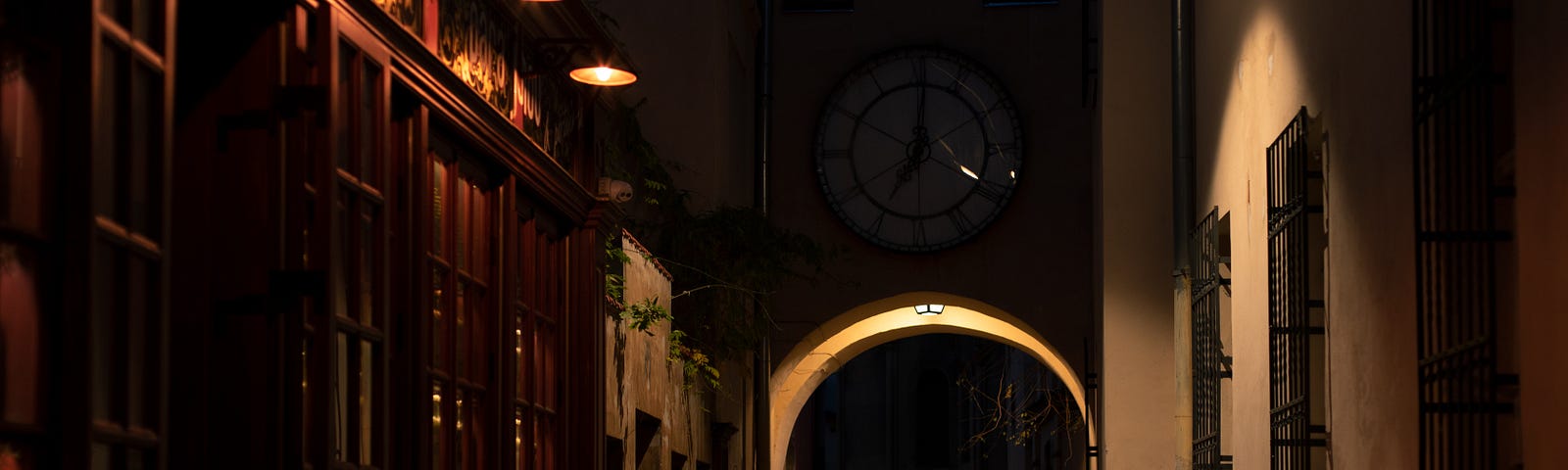 A person in a black suit is seen walking in a dimly lit cobblestone alley. A big clock is over an arch that the person is heading towards.