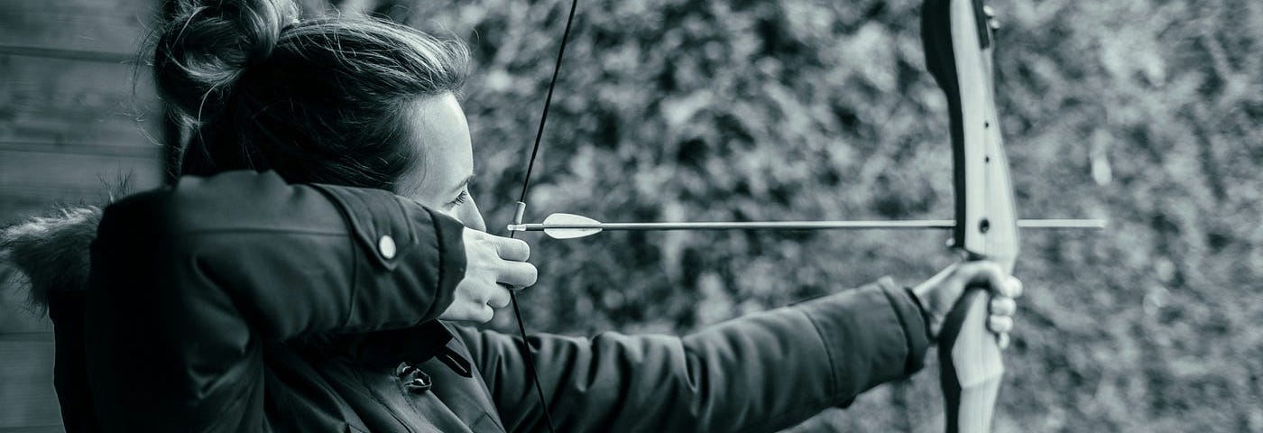 A woman holds a bow and an arrow and aims at an invisible target in this black and-white photo.