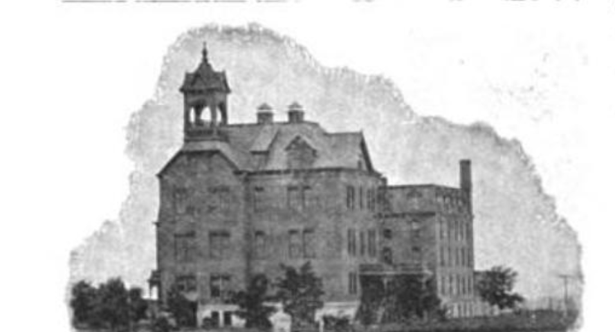 A large 4 story brick building with a cupola and small trees around the sides of the building that are visible.