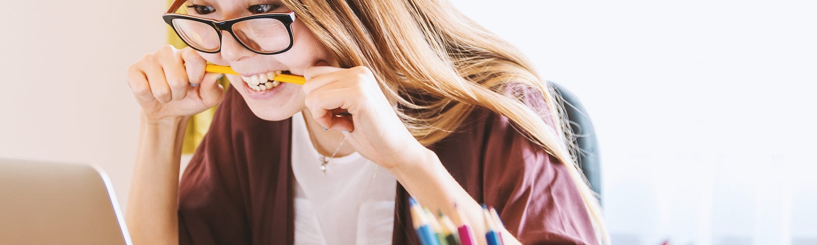 A lady in glasses sits at a desk over a laptop. She looks excited and holds a yellow pencil in her mouth with both hands. On her desk is a pen holder containing an assortment of coloured pencils.