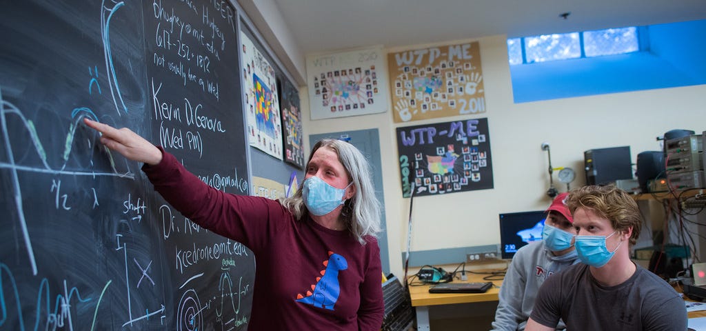 A woman wearing a mask stands at a blackboard explaining a mathemetical concept to two masked, male students