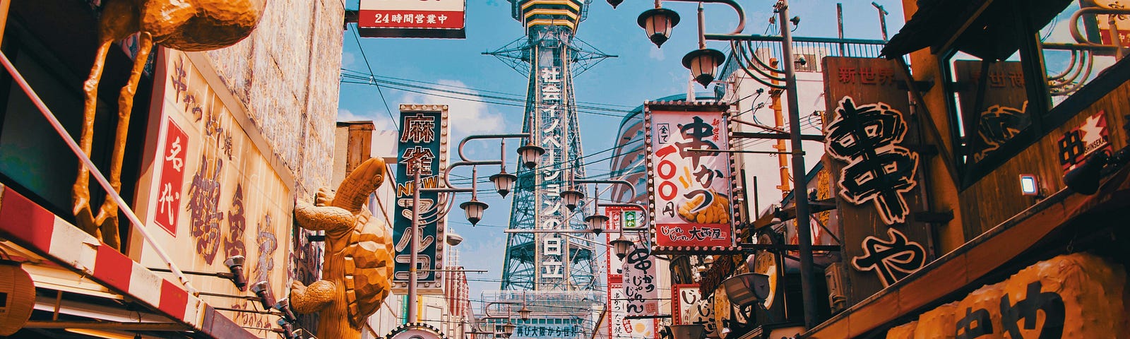 Streetscape of Osaka, Japan, on a sunny day. The photo has a grainy, film-like quality. The blue and white Tsutenkaku Tower is at the back of the photo in the centre. Shops with signs and 3-D characters on them line the street leading to the tower.
