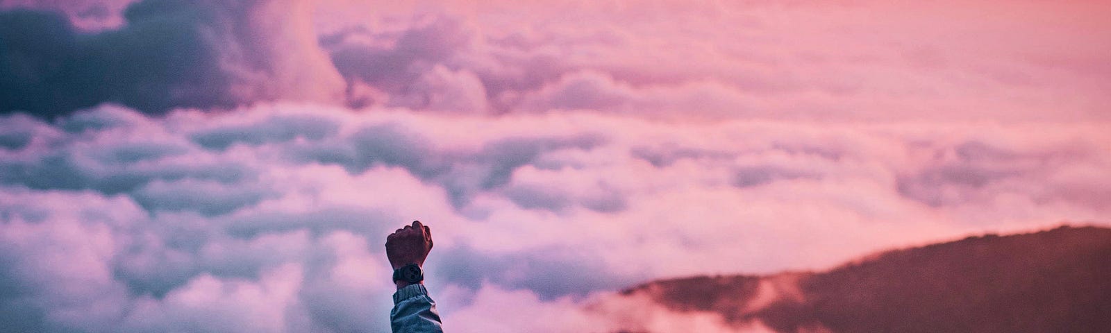 Young man sitting on top of mountain above the clouds watching the sun rise. His left hand is raised, celebrating the glory of it all.