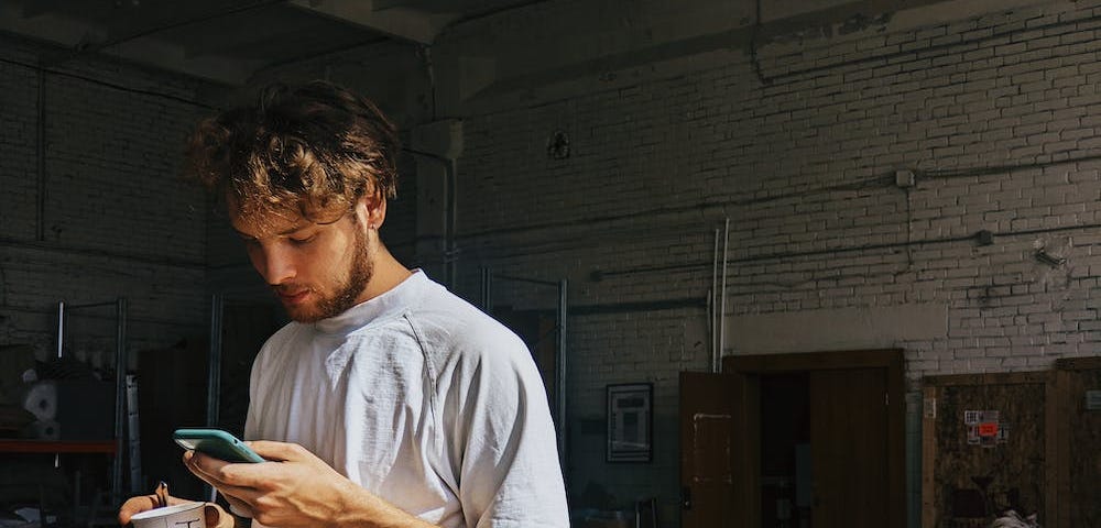 A young man is holding a paper cup while surfing social media on his phone. // onetoforseeforall/Pexels.