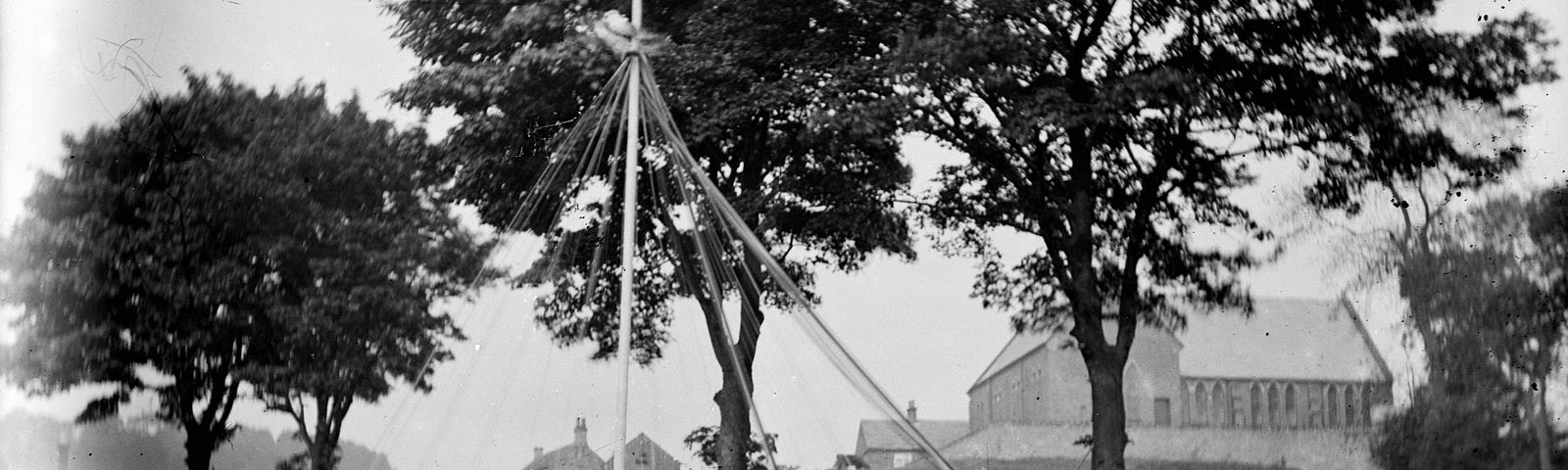 a black and white photo showing girls dressed in white standing around a maypole with trees in the background