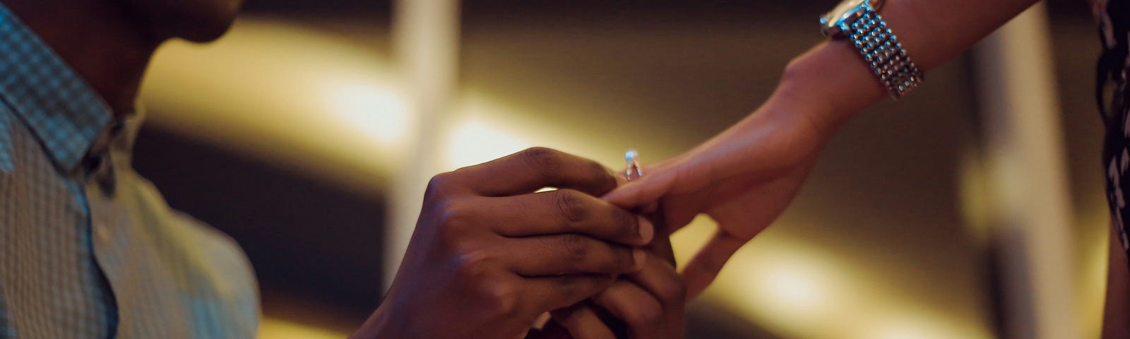 A close up, color photograph that partially shows a kneeling man, placing a ring on a woman’s hand.