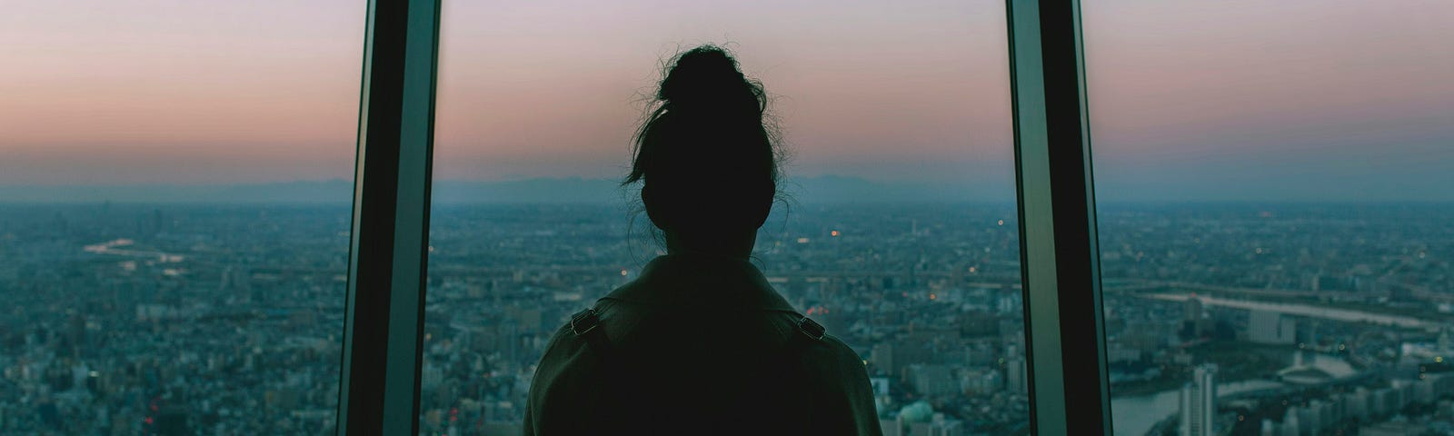 photograph of someone wearing a backpack and their hair in a topknot looking out from an upper airport or hotel window across a vast city in the twilight