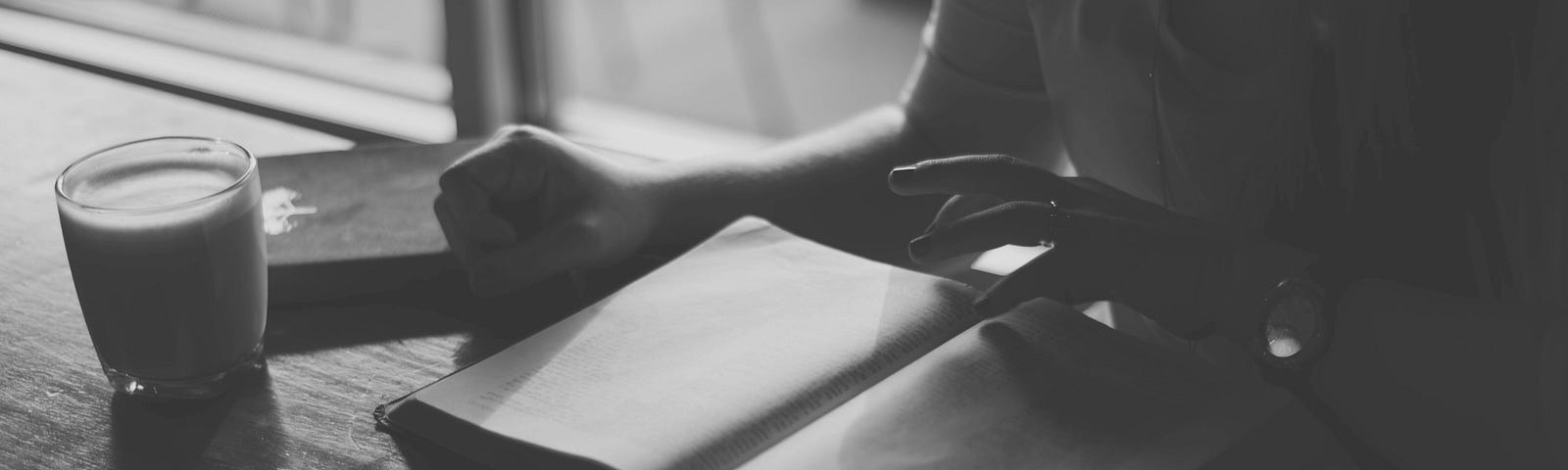 black and white photo showing a book open on a table beside a glass of drink and the torso of a person reading the book