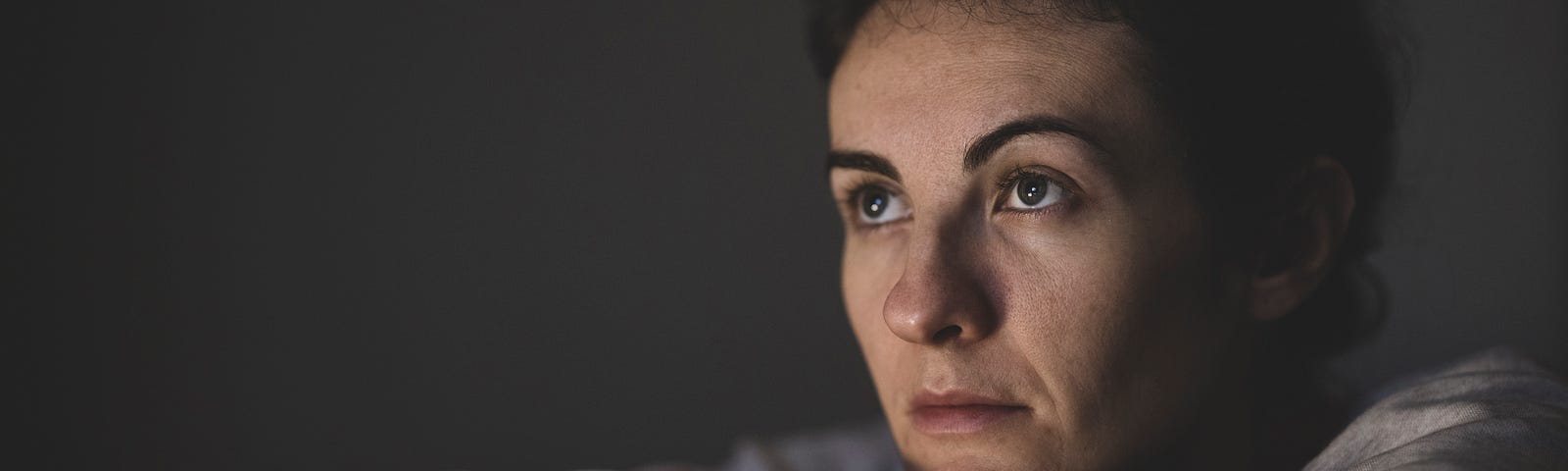 A sitting woman wearing a grey sweatshirt and fuzzy pajama pants holds her knees to her chest, looking exhausted and worried. The foreground is dark, indicating it is late at night.