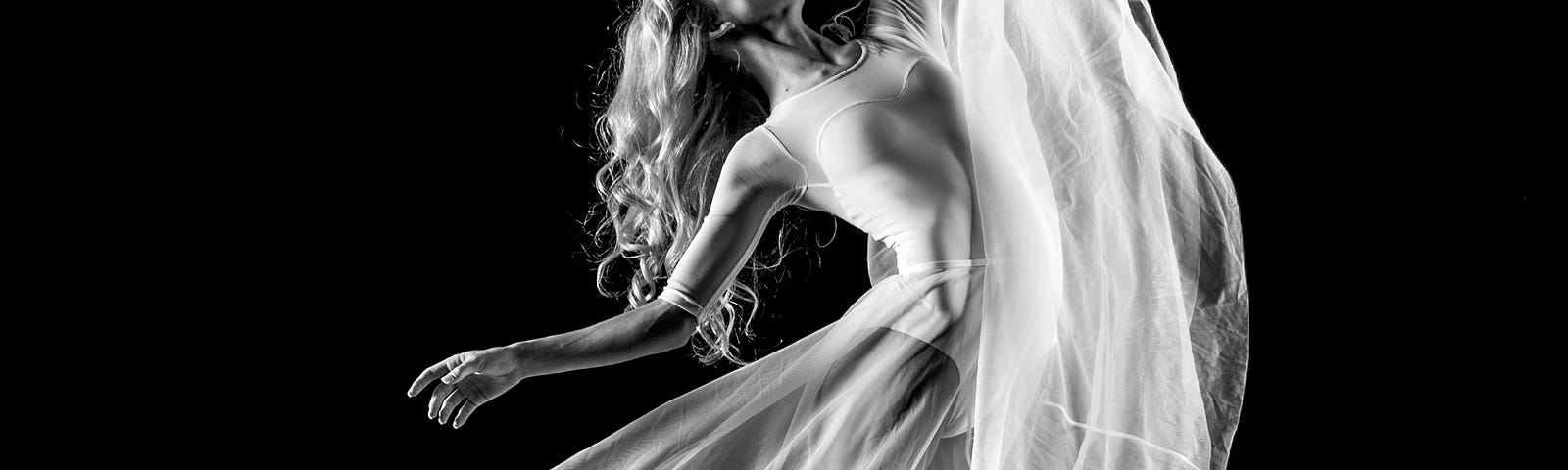 Black and white picture of a woman with long blonde hair, sheer fitted leotard and mesh flowing long skirt, dancing in her bare feet.