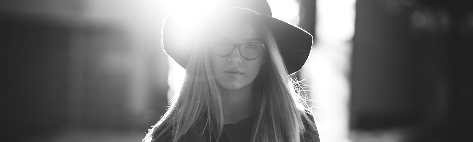 Young girl with long blond hair in a black and white photo. She is wearing glasses and a hat and is looking straight at the camera solemnly.