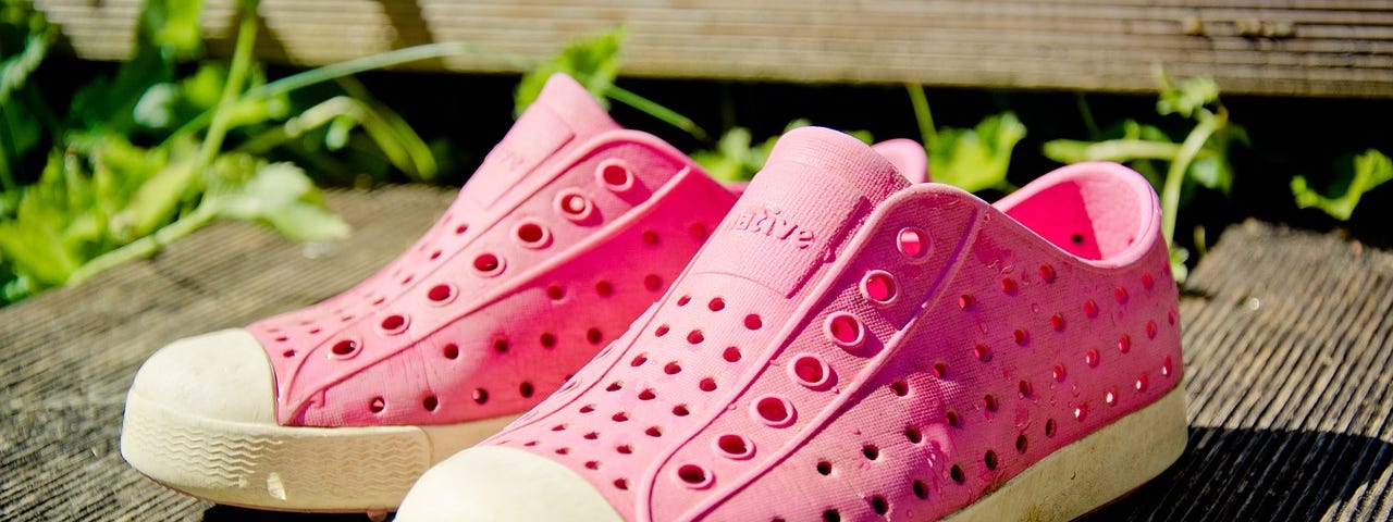 two pink “crocs” shoes on a porch in the sun.