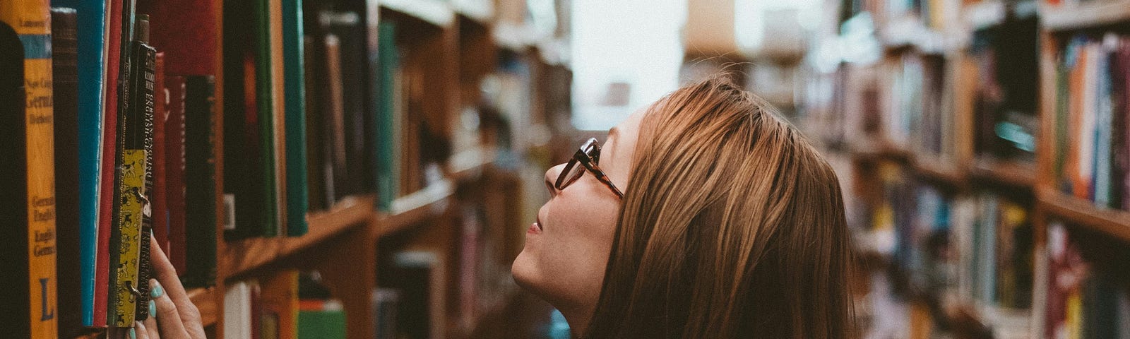 A young college student browsing the wooden, bookshelves of a college library.