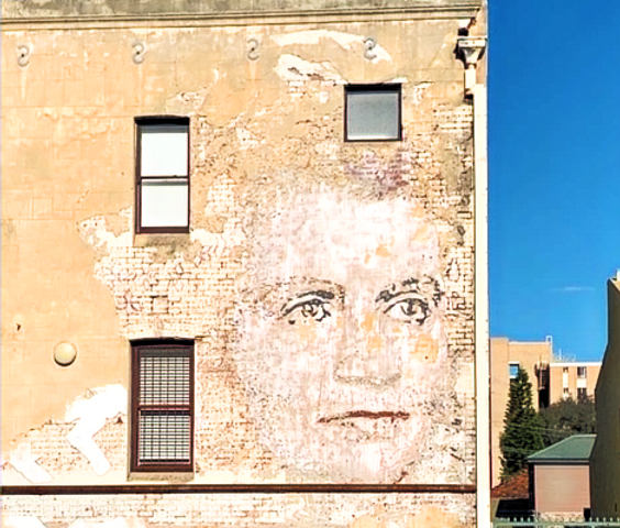 Street view of a giant, fading portrait of a woman’s face on a hotel wall. Pale yellow brickwork surrounds it.