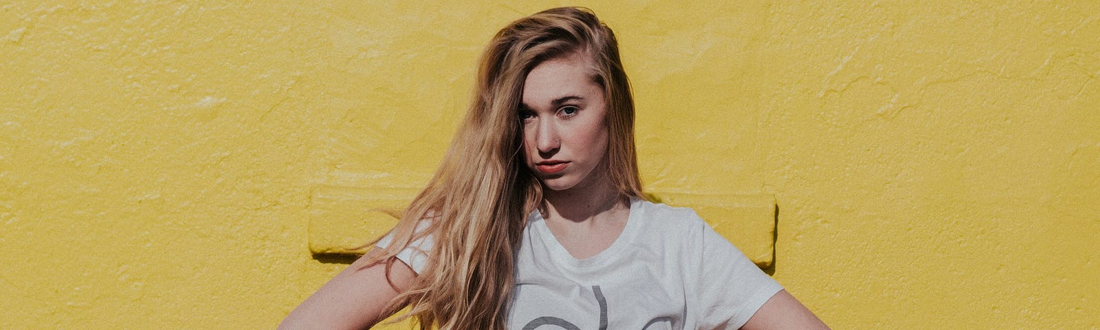 A white woman wearing black jeans and a white shirt, with long blonde hair swept over her shoulder, stands with her hands on her hips, in front of a yellow wall.