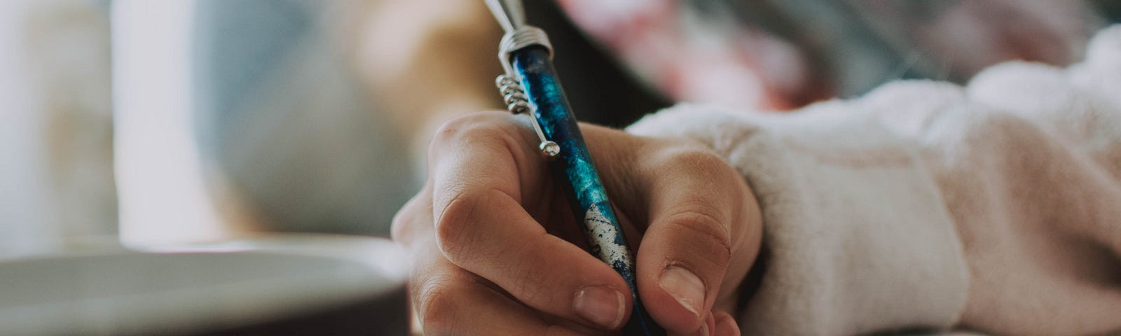 Um braço encasacado de uma mulher escreve em um caderno com uma caneta azul. Na frente do caderno, há uma xícara e um óculos.