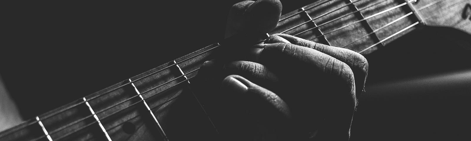 This black-and-white photo shows the neck of an acoustic guitar being played with a hand forming a chord in the 4th fret down.