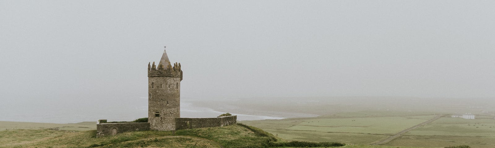 A small stone tower sits on a hill on a coast. The sky is foggy.